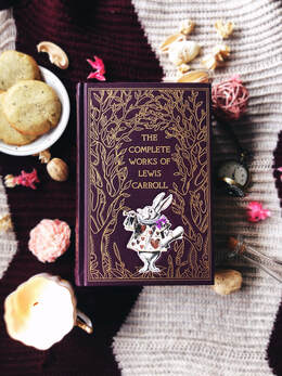 A purple copy of Lewis Carroll's works with the white rabbit on the front sits on a purple and white blanket background with a tea cup candle and flowers around it. 