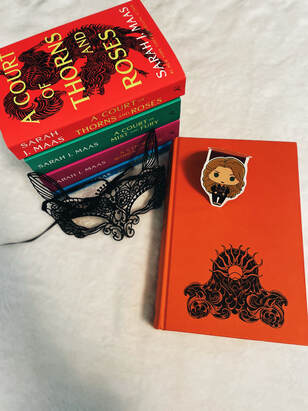 A stack of the Court of Thorns and Roses series with book one on the top sitting behind a black lace cat mask next to the newest book of the series on a white blanket background. On top of Court of Silver Flames sits a bookmark of Sarah J Maas holding a copy of the book. 