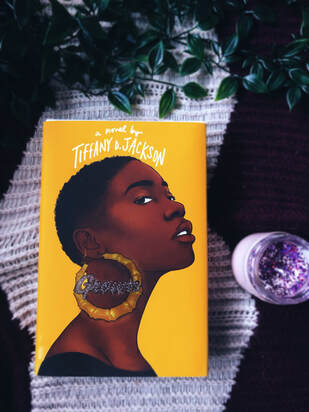 A copy of Grown by Tiffany Jackson sits on a purple and white blanket background with green ivy above it and a glitter purple candle next to it. 