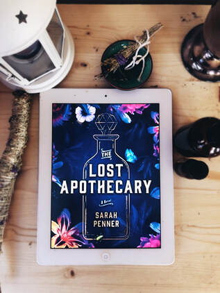 A copy of The Lost Apothecary on an ereader sits on a light wooden table background with sage and essential oils around it. 