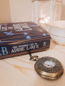 The bottom half of a copy of Addie LaRue sits on a light wooden table with a pocket watch, a lit candle and a tea cup. 