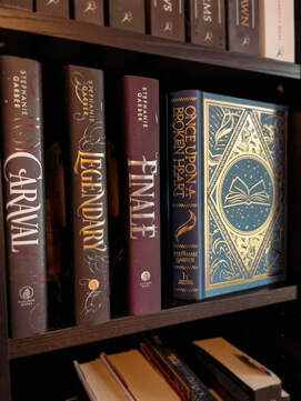 Copies of Caraval, Legendary, and Finale lead up to the blue and gold cover for Once Upon A Broken Heart on a black book shelf. 