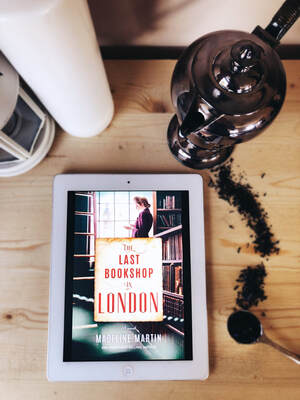 Ipad with The Last Bookshop in London cover displayed showing a woman at the window of an old bookstore. Ipad sits on a light wooden table with two candles and a teapot that has tea grounds spilling from the pot. 