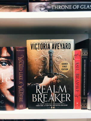 A copy of Realm Breaker by Victoria Aveyard sits among other books on a book shelf. The cover is gold with a bleeding hand holding a sword. 
