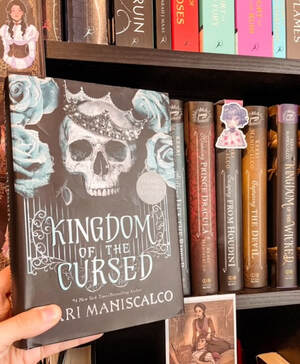 A black and blue copy of Kingdom of the Cursed is held in front of a book shelf with the rest of Kerri Maniscalco's books. The cover has blue roses on a black background with a white skill in the middle wearing a silver crown. 
